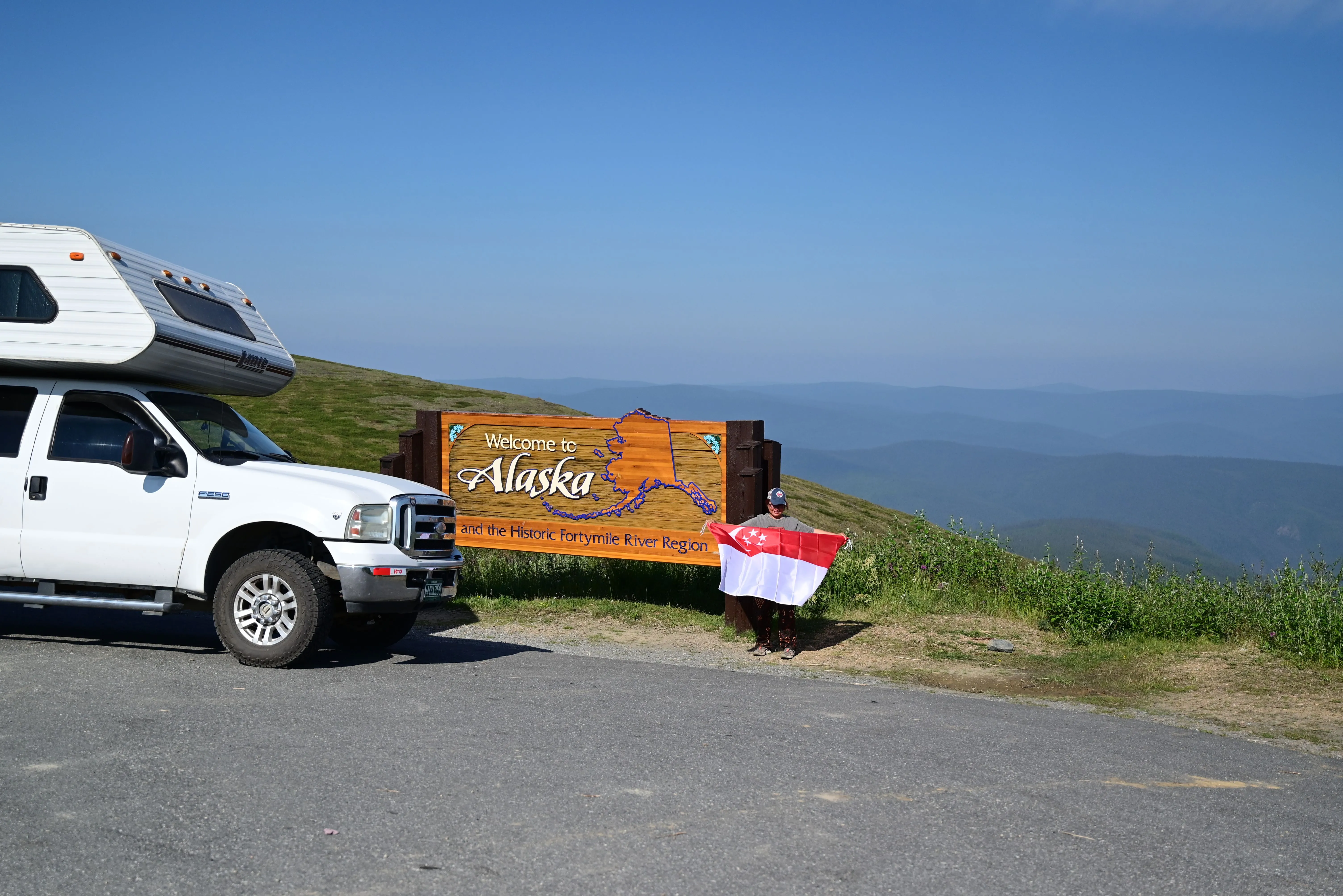 A woman holding up the Singapore flag infront of a Welcome to Alaska sign